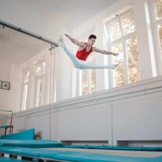 Akrobat mit rotem Top und weißer Hose im Spagat in der Luft bei Uni Sportkurs