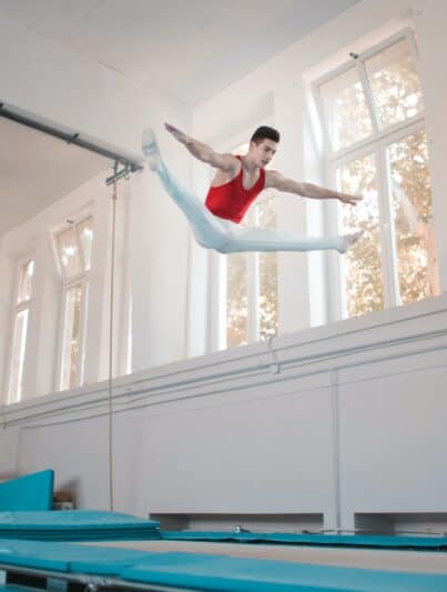 Akrobat mit rotem Top und weißer Hose im Spagat in der Luft bei Uni Sportkurs