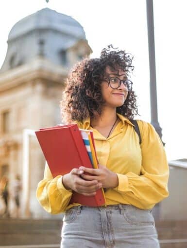 Frau in gelber Jacke, die vor historischem Gebäude rotes Buch hält