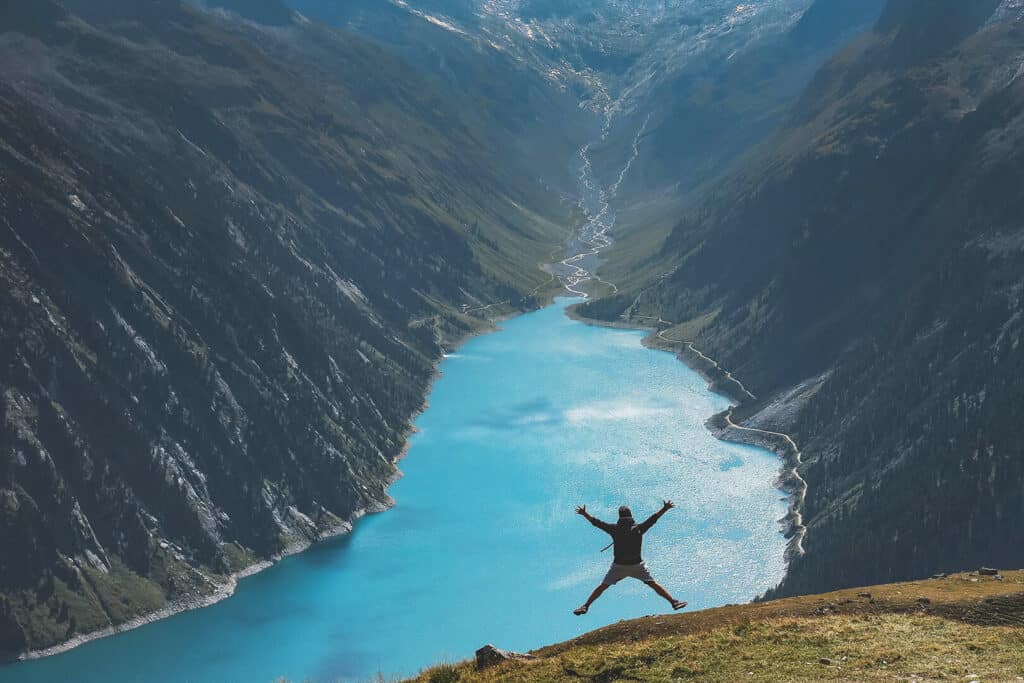 Bergsee vom Berg aus fotografiert mit einem Menschen der vor Freude springt, weil er eine klimafreundliche Reise macht. 