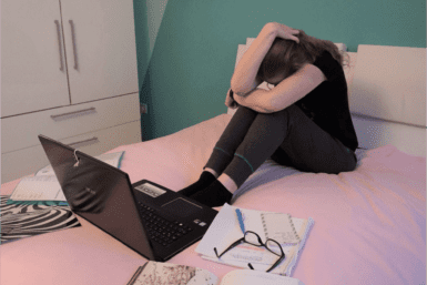 Frau die mit Laptop und Lernunterlagen verzweifelt auf einem Bett sitzt
