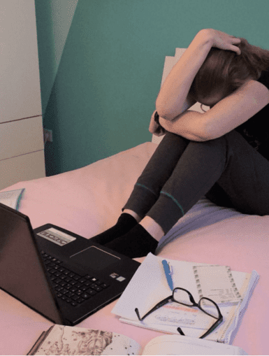 Frau die mit Laptop und Lernunterlagen verzweifelt auf einem Bett sitzt