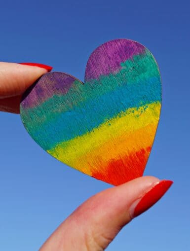 Eine Hand hält ein Herz aus Papier, das in Regenbogenfarben angemalt ist.