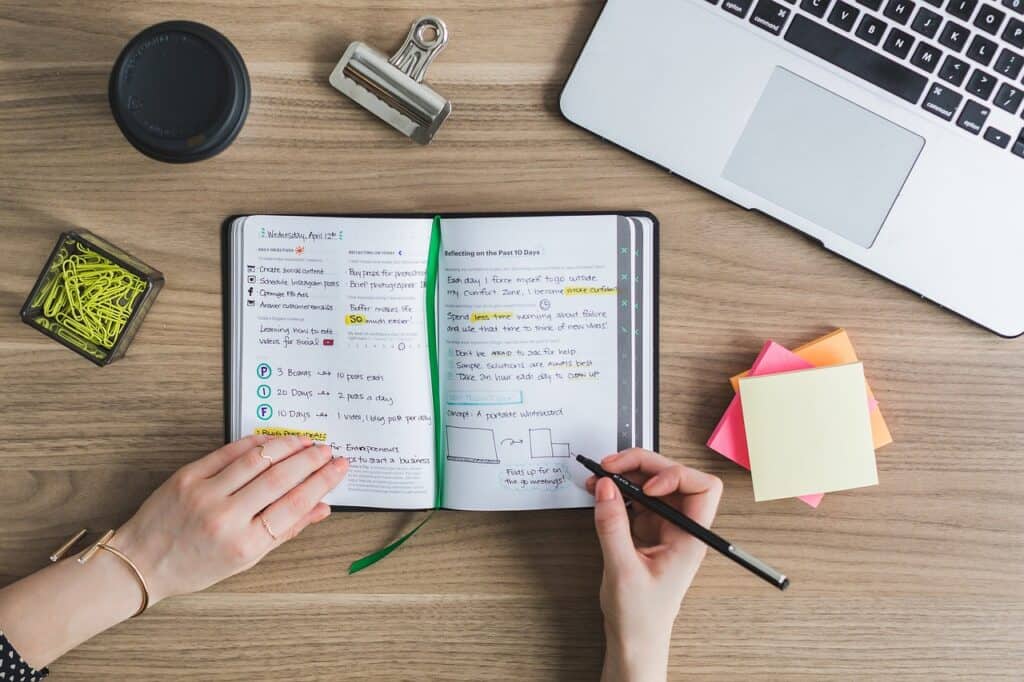 Person schreibt Informationen zum Lernen in ein Notizbuch. Daneben liegen ein Stapel Post-its, grüne Büroklammern in einer Dose, ein schwarzer Coffee-to-go-Becher und ein Laptop. 