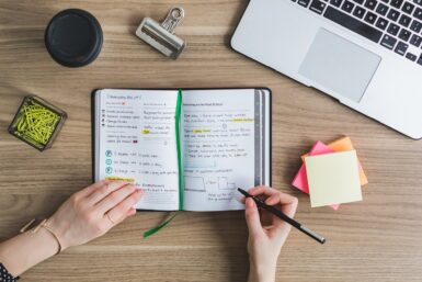 Person schreibt Informationen zum Lernen in ein Notizbuch. Daneben liegen ein Stapel Post-its, grüne Büroklammern in einer Dose, ein schwarzer Coffee-to-go-Becher und ein Laptop.