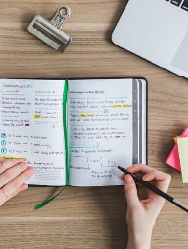 Person schreibt Informationen zum Lernen in ein Notizbuch. Daneben liegen ein Stapel Post-its, grüne Büroklammern in einer Dose, ein schwarzer Coffee-to-go-Becher und ein Laptop.