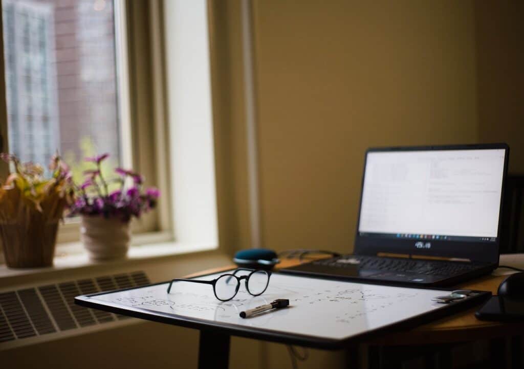 Ein Laptop steht auf einem Tisch. Vor dem Laptop liegt ein Whiteboard mit einem Whiteboardmarker und einer Brille darauf.
