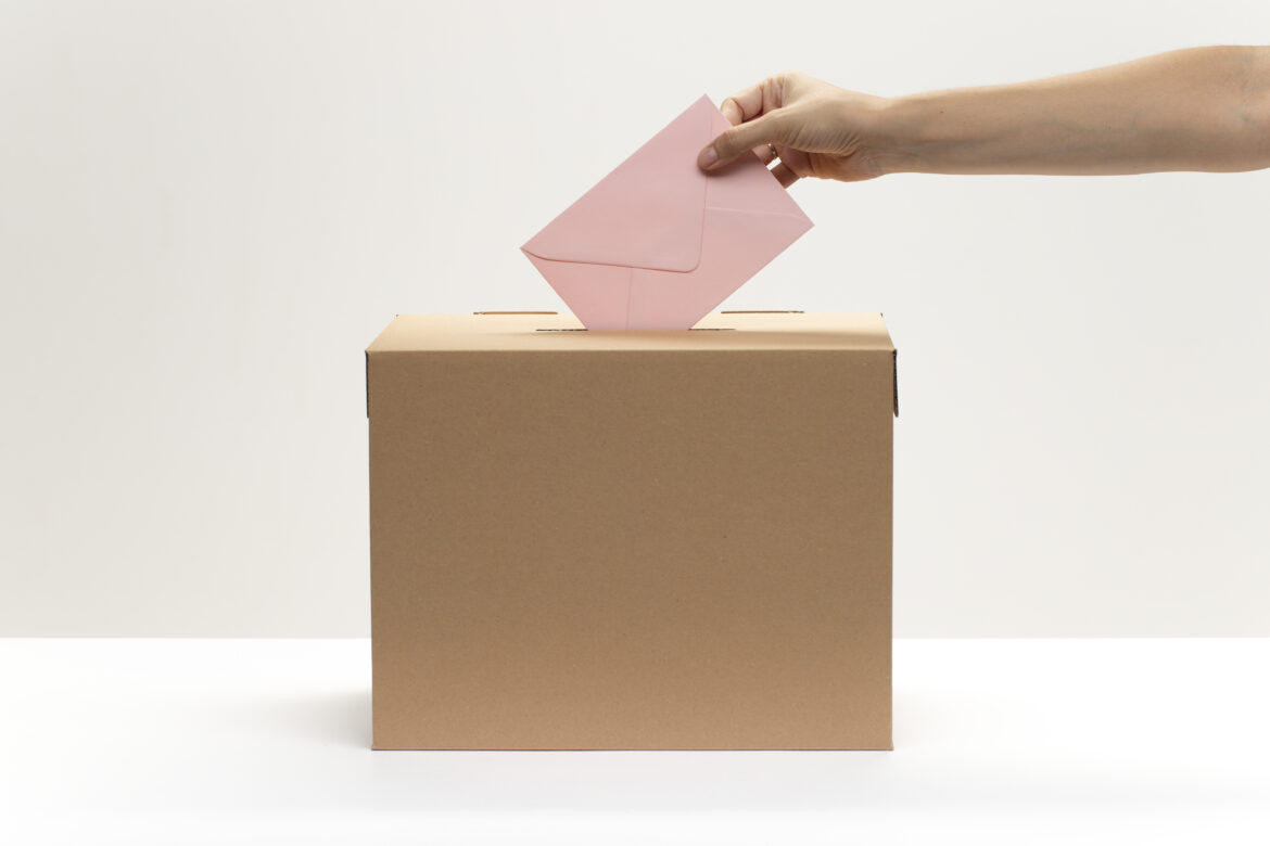 Eine Hand wirft bei einer Wahl einen rosanen Stimmzettel in eine braune Wahlurne. Der Bildhintergrund ist weiß.