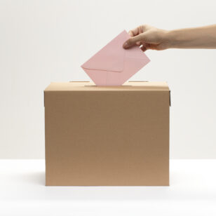Eine Hand wirft bei einer Wahl einen rosanen Stimmzettel in eine braune Wahlurne. Der Bildhintergrund ist weiß.