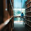 Man blickt durch einen Gang von Bücherregalen. Am Ende des Gangs sitzt eine Person vor einem Tisch und liest Bücher. Der Tisch ist vor einer Fensterfront.