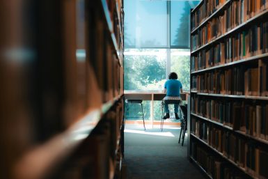 Man blickt durch einen Gang von Bücherregalen. Am Ende des Gangs sitzt eine Person vor einem Tisch und liest Bücher. Der Tisch ist vor einer Fensterfront.