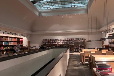 Zu sehen ist eine Bibliothek von innen. An den Wänden sind Bücher in Regalen einsortiert. Die Decke ist aus Glas. In Mitten der Bibliothek ist ein Geländer. Man kann hier in den unteren Stock sehen. (c) Shona Stern