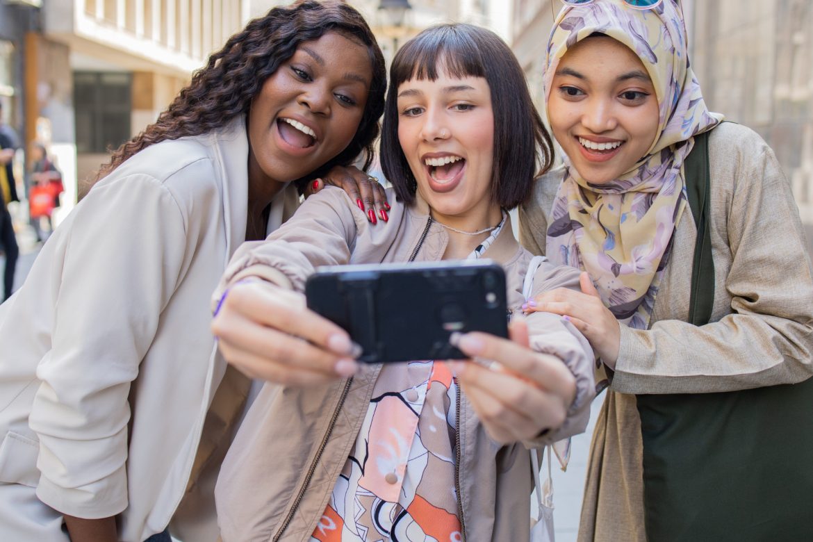 Drei junge Frauen werden von vorne gezeigt, wie sie sich hinter einem Handy zusammenlehnen, um ein Selfie zu machen. Copyright: Getty Images