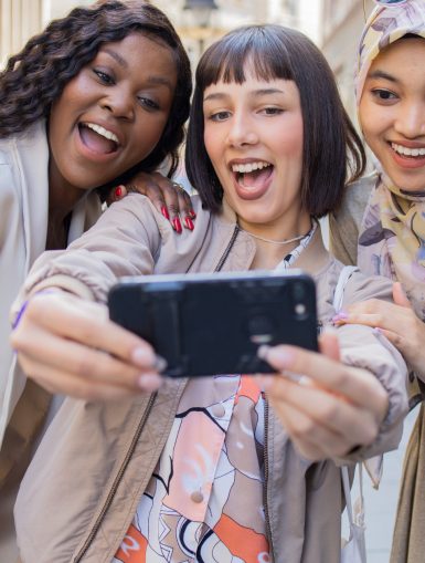 Drei junge Frauen werden von vorne gezeigt, wie sie sich hinter einem Handy zusammenlehnen, um ein Selfie zu machen. Copyright: Getty Images